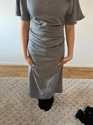 Grå midiklänning med rynkade detaljer - Stilren grå klänning i midi-längd med rynkade detaljer i midjan och korta ärmar. Klänningen har en rak passform och slits bak för extra rörelsefrihet. Tillverkad i ett mjukt tyg som ger en elegant känsla.