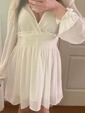 Vit omlottklänning med lång ärm - Säljer en söt vit kortklänning med omlottdesign och smockad midja. Klänningen har långa, luftiga ärmar med resår vid ärmslut och ett flowy fall. Perfekt för sommarens alla tillfällen, även student och skolavslutning! 😊 Kan skickas snabbt!