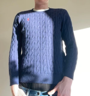 Mörkblå kabelstickad tröja polo ralphlauren - Snygg mörkblå kabelstickad tröja från Polo Ralphlauren med rund halsringning och diskret röd logga broderad på bröstet. Tröjan har ribbade muddar vid ärmslut och nederkant. Perfekt för lager på lager och klassisk stil.