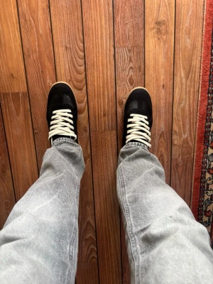 Svarta sneakers med brun sula - Snygga svarta sneakers med kontrasterande vita skosnören och beige gummisula. Klassisk låg modell med stilren design och paneler i mocka och textil. Perfekta för dig som vill ha en clean och tidlös look till jeans eller streetwear.