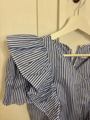 Blåvit randig blus med volanger - Snygg blåvit randig blus med korta ärmar och stora volangdetaljer framtill. Blusen har en v-ringning och är tillverkad i ett lätt bomullsliknande material.storlek 11-12 år (152)