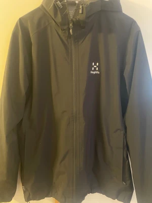 Svart vindjacka från Haglöfs - Snygg svart vindjacka från Haglöfs med dragkedja framtill och justerbar huva. Jackan har minimalistisk design med logga på bröstet och är tillverkad i ett lätt, vattenavvisande material. Perfekt för blåsiga dagar och outdoor-aktiviteter.