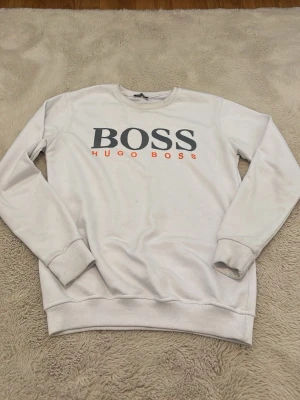 Tröja - Säljer en stilren vit sweatshirt från Hugo Boss med stor logga i svart och orange text på bröstet. Tröjan har rund hals, långa ärmar och ribbade muddar vid ärmslut och nederkant. Perfekt för en avslappnad och trendig look.