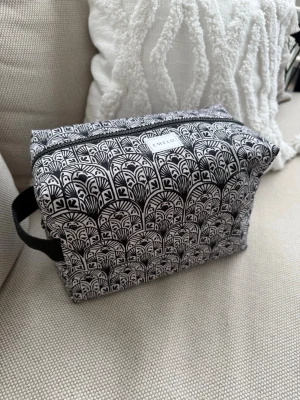 Svartvit necessär från Emelo - Necessär i tyg från Emelo med svartvitt grafiskt mönster och dragkedja upptill. Praktiskt handtag på sidan och rektangulär form. Svart insida med innerficka. Perfekt för smink eller småprylar. Stilren och enkel att ta med sig. Storlek large. Mått ca 27x17x12cm