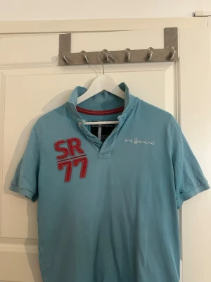 Ljusblå Sail Racing skjorta - Ljusblå kortärmad skjorta från Sail Racing med SR77-brodyr i rött och vitt på bröstet. Skjortan har en unik krage med knappdetalj och diskret logga på ryggen. Perfekt för en avslappnad och sportig stil.