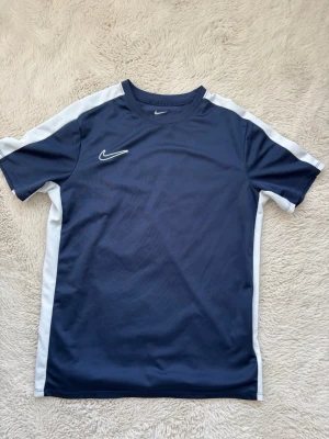 Blå Nike Dri-FIT t-shirt XL - Säljer en blå Nike Dri-FIT t-shirt med vita detaljer längs sidorna och axlarna. Perfekt för träning eller sport, tillverkad i ett lätt och ventilerande material som håller dig torr. Klassisk Nike-logga på bröstet och halsen. Passar dig som gillar sportiga kläder.