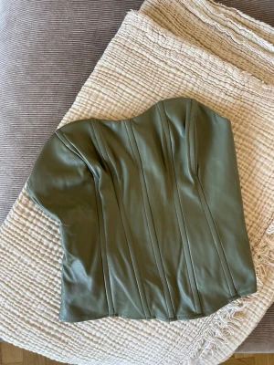 Olivgrön korsett i skinnimitation - Snygg olivgrön korsett med markerade sömmar och formad överkant. En kortare variant av en korsett!! Aldrig använd men prislappen är borttagen💚