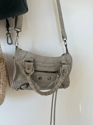 Grå handväska - Säljer en grå handväska i mjukt läder med både axelrem och flätat handtag. Väskan har en framficka med dragkedja, dekorativa nitar och långa läderdetaljer. Perfekt för dig som gillar en cool och tidlös look. Köptes för 800 kr
