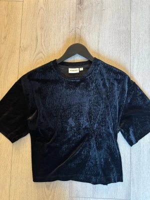 Svart sammetst-shirt från Kappahl - Mörk, svart t-shirt i mjuk sammet från Kappahl. Modellen är croppad med korta ärmar och rund halsringning. Perfekt för dig som gillar en stilren men ändå lyxig vibe. Passar till många olika looks och är superbekväm.
