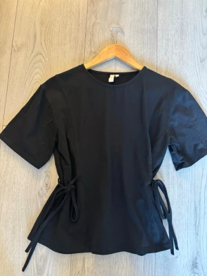 Svart topp med knyt i midjan - Snygg svart topp med korta ärmar och rund halsringning. Toppen har knytband i midjan på båda sidor som ger en cool och justerbar passform. Perfekt till jeans eller kjol för en clean look.