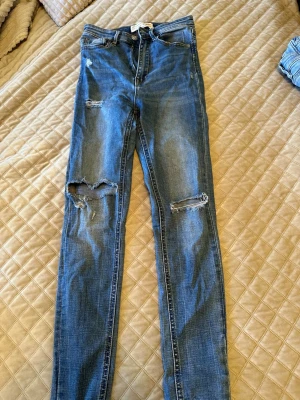  jeans Stradivarius - Säljer ett par blå super high waist jeans från Stradivarius i storlek 34. Jeansen har skinny fit, slitningar och hål på benen. 