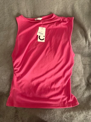 Rosa ärmlös topp från Cubus -  En helt ny snygg rosa ärmlös topp från Cubus i mjukt och stretchigt material.  Den är aldrig använd! På ena axeln har den en scrunch. Toppen har rund halsringning och en clean, enkel design som passar perfekt till jeans eller kjol. Perfekt för dig som gillar färg och vill ha en bekväm topp till vardagslooken.