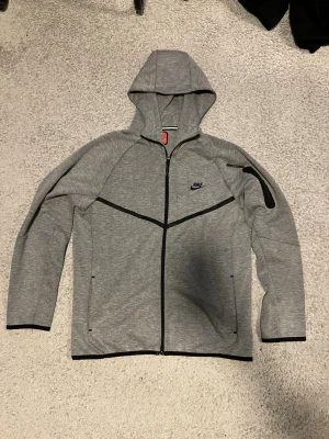 Grå Nike Tech Fleece hoodie - Snygg grå Nike Tech Fleece hoodie med hel dragkedja och svarta detaljer. Har huva, två sidofickor med dragkedja och en extra ficka på ärmen. Tillverkad i mjukt och lätt material som ger en sportig look. Perfekt för dig som gillar streetwear och sportig stil.