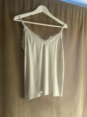 Vit spetsdetaljerad linnetopp Amisu - Säljer en vit linnetopp från Amisu med tunna axelband och v-ringad halsringning dekorerad med spets. Toppen har en enkel och stilren design, perfekt att matcha med jeans eller kjol. Materialet känns mjukt och lätt.