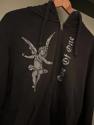 Svart hoodie One Of One med strassängel - Svart hoodie från One Of One i storlek medium med dragkedja, huva och snörning. På framsidan finns en stor strassängel och texten 'One Of One' i glittrande stenar. Hoodien har känguruficka och är gjord i mjukt bomullsmaterial.