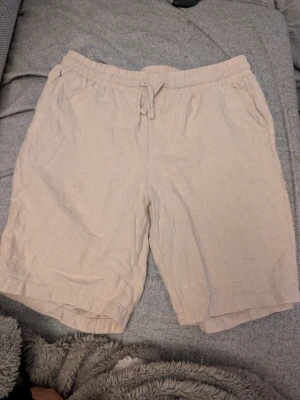 Beige linneshorts med snörning stl 174 - Snygga beige shorts i linne med elastisk midja och snörning framtill. Perfekta för varma dagar och har en avslappnad passform med fickor på sidorna. Lätta att matcha med olika toppar för en chill look. Mycket fint t skick