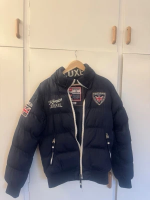 Mörkblå pufferjacka från Deeluxe - Snygg mörkblå pufferjacka från Deeluxe med broderade patchar och Union Jack-detaljer på ärmen och bröstet. Jackan har hög krage, vita dragkedjor och två framfickor med dragkedja. Perfekt för kalla vinterdagar och riktigt street-inspirerad stil.