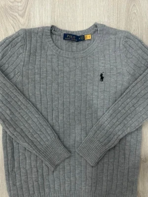Grå kabelstickad tröja Polo Ralph Lauren - Kabelstickad grå tröja från Polo Ralph Lauren med rund halsringning och svart broderad logga på bröstet. Tröjan har långa ärmar och ribbade muddar vid ärmslut och nederkant. Perfekt för en stilren och klassisk look.