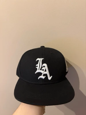 Svart LA keps med brodyr - Säljer en svart keps med rak skärm och vit broderad LA-logga framtill. På sidan finns en rund brodyr med texten 'Los Angeles California' och en jordglob. Klassisk streetwear-stil, perfekt för dig som gillar sportiga accessoarer.
