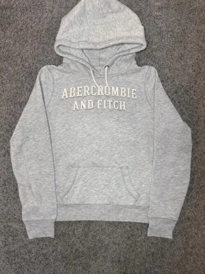 Grå hoodie från Abercrombie & Fitch - Snygg ljusgrå hoodie från Abercrombie & Fitch med klassisk logga broderad på bröstet. Hoodien har huva med dragsko, magficka och ribbade muddar vid ärmar och nederkant. Perfekt för en avslappnad och trendig look.