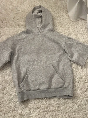 Grå hoodie med korta ärmar - Säljer en stilren grå hoodie med korta ärmar och klassisk magficka. Hoodien har ribbade muddar nedtill och en mjuk huva. Klippt hemma själv 
