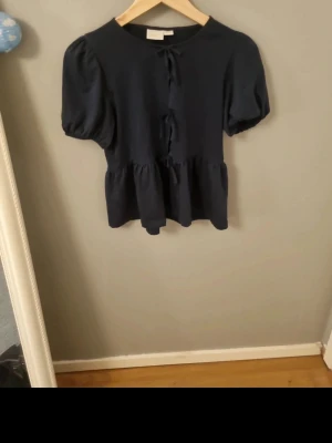 T-shirt från gina trico  - Trendig svart blus med puffärmar och tre dekorativa knyt framtill. Blusen har en lätt volang nedtill och är tillverkad i ett mjukt tyg som känns skönt mot huden. Perfekt för dig som gillar en söt och stilren look. Använd fåtal gånger och är sågott som ny! Är i storleken 164 och finns ej i butik.