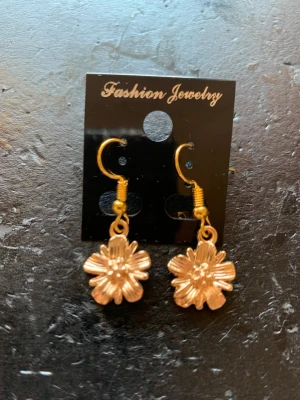 Guldiga blommiga örhängen - Snygga örhängen i guldfärgad metall med blommor som hänger ner. De har en krok för enkel användning och en cool, glansig finish. Perfekta för dig som vill ha en söt och trendig accessoar med blommig touch.