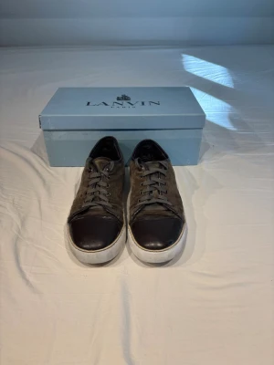 Bruna Lanvin sneakers i mocka och läder - Snygga sneakers från Lanvin i brunt mocka och mörkbrunt läder på tån. Skorna har vita sulor och klassisk snörning. Perfekta för dig som gillar exklusiva och stilrena sneakers med en lyxig känsla.