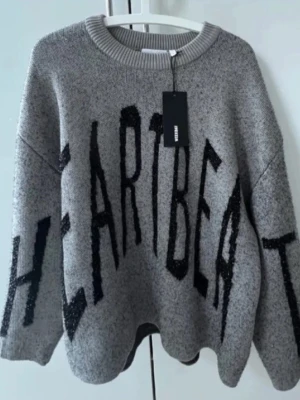 Grå oversized tröja - Snygg grå stickad tröja från weekday med stor svart text 'HEARTBEAT' över hela framsidan. Tröjan har rund halsringning, avslappnad passform och ribbade muddar vid halsen. Perfekt för dig som gillar statement-plagg och vill ha en trendig look 🥰