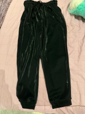 Svarta mjukisbyxor i velour - Säljer ett par svarta mjukisbyxor i glansigt velourmaterial med resår i midjan och muddar vid bensluten. Byxorna har en avslappnad passform och dragsko i midjan för extra komfort. Perfekta för chill dagar eller när du vill vara extra bekväm.