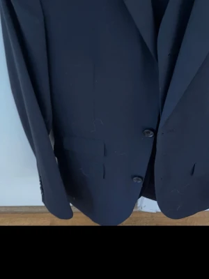 Mörkblå kavaj från Oscar Jacobson - Snygg mörkblå kavaj från Oscar Jacobson med klassisk passform och två knappar framtill. Kavajen har innerfoder och flera fickor, perfekt för en stilren look. Materialet är ullblandning som ger en exklusiv känsla.