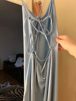 Ljusblå långklänning med snörning - Snygg ljusblå långklänning med öppen rygg och dekorativ snörning baktill. Klänningen har en slits på sidan och är tillverkad i ett mjukt, tunt material som ger ett elegant fall. Perfekt för dig som vill sticka ut med en trendig och stilren look.