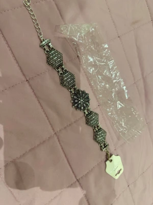 Silvrigt armband med vintagekänsla - Unikt silvrigt armband med detaljerade geometriska länkar och en blomformad mittdel. Armbandet har små gnistrande stenar som ger extra bling och en justerbar kedja för perfekt passform. Perfekt accessoar för dig som gillar retrostil och statement-smycken.