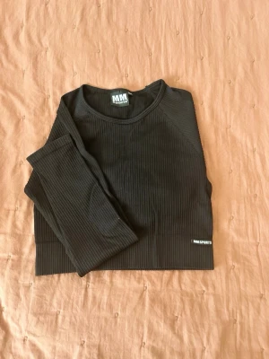 Svart ribbad crop top MM Sports - Säljer en svart ribbad crop top från MM Sports med lång ärm och rund hals. Tröjan har en tight passform och är tillverkad i stretchigt material som passar perfekt till träning eller gym. Diskret MM Sports-logga nertill.