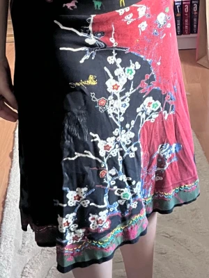 Desigual kjol - Kjol från Desigual med blommigt mönster i vitt, grönt och rött på svart och röd bakgrund. Kjolen har en färgglad kant nertill och är tillverkad i 100% mjuk bomull.  Kjolen är stretchig i midjan och kan användas både högmidjat och lågmidjat och den passar storlek M/L samt XL då den har en bekväm och stretchig passform.  Jättefint mönster och väldigt bra skick utan några skador eller fläckar!