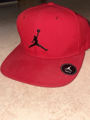 Röd Jordan keps med broderad logga - Snygg röd keps från Jordan med den klassiska svarta Jumpman-loggan broderad framtill. Kepsen har platt skärm och justerbar snapback baktill för perfekt passform. Tillverkad i tåligt bomullsmaterial och passar ungdomar.