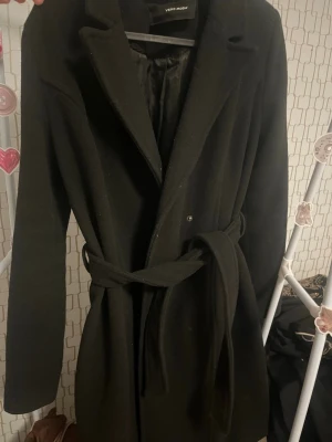 Svart rock från Vero Moda med skärp - Stilren svart kappa från Vero Moda med klassisk krage och knytband i midjan. Rocken har en slät yta och är helfodrad med svart innerfoder. Inga defekter. 