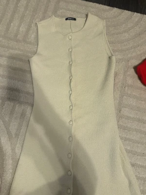 Beige ärmlös klänning Gina Tricot - Snygg beige ärmlös klänning från Gina Tricot med knappar längs hela framsidan. Klänningen har en enkel, rak passform och är tillverkad i ett mjukt, stickat material. Perfekt för dig som gillar stilrena och bekväma plagg. Köpt för 399kr