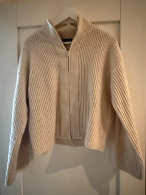Beige stickad ziptröja från 157 - Säljer en beige stickad tröja från 157 med dragkedja framtill och hög krage. Tröjan har breda ärmar och ribbad struktur som ger en snygg och avslappnad look. Perfekt att slänga på sig när det är kyligt ute.