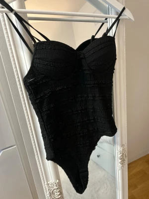 Svart spetsig body med vadderade kupor - Snygg svart body med tunna axelband och vadderade kupor för extra stöd. Bodyn har ett spetsigt mönster och fina detaljer som ger en trendig look. Perfekt att styla under en skjorta eller kavaj. Materialet är mjukt och följsamt.