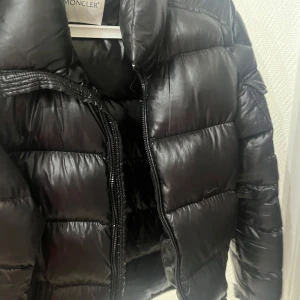 Svart dunjacka från Moncler - Snygg svart dunjacka från Moncler med glansig finish och klassisk quiltad design. Jackan har hög krage, dragkedja framtill och Moncler-logga på ärmen. Perfekt för kalla vinterdagar och riktigt varm tack vare dunfyllningen.