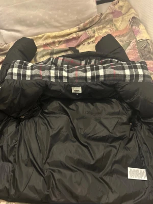 Svart dunjacka från Burberry - Snygg svart dunjacka från Burberry med klassiskt rutigt foder i svart, vitt och rött. Jackan har huva, dragkedja och tryckknappar samt en patch med 'Burberry London England' på ärmen. Perfekt för kalla vinterdagar.