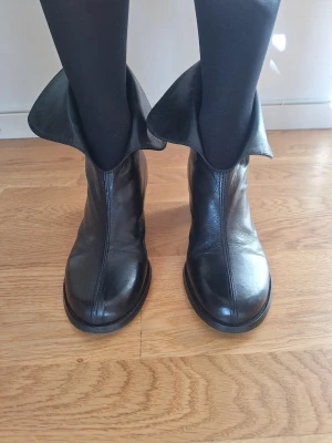 Svarta läderstövletter med uppvikt kant - Säljer ett par svarta stövletter i äkta läder med unik uppvikt kant upptill. Skorna har rundad tå och en stilren, minimalistisk design utan synliga detaljer eller dekorationer. Perfekta för dig som gillar en edgy och modern look.