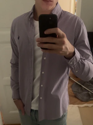 Lila rutig skjorta från Ralph Lauren - Säljer en lila och vit rutig skjorta från Ralph Lauren med klassisk button-down krage och broderad logga på bröstet. Skjortan har lång ärm, vita knappar och är tillverkad i mjuk bomull. Perfekt för en clean och stilren look. Bröstmått: 47 cm Längd: 70 cm