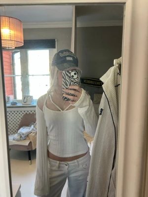 Vit ribbad topp med unika band - Trendig vit topp med ribbad struktur och unika dubbla axelband som korsar framtill. Toppen har långa ärmar och en mjuk, stretchig känsla. Perfekt för dig som gillar en stilren men ändå cool look.