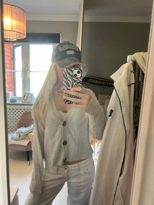 Beige cropped kofta med knappar - Säljer en beige cropped kofta med stora bruna knappar och v-ringning. Koftan har lång ärm, ribbade muddar och är gjord i ett mjukt, stickat material. Perfekt att styla med jeans eller kjol för en chill look.