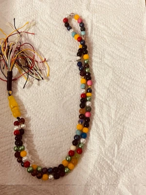 Färgglad handgjord bönkedja - Unik bönkedja med pärlor i flera färger som rött, gult, grönt, blått och vitt. Kedjan har en tofs i ena änden med trådar i olika färger och pärlor av plast. Perfekt som accessoar eller dekoration. Ingen synlig märkning.