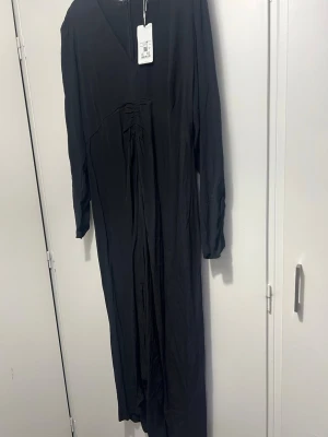 Svart långklänning med v-ringning - Stilren svart långklänning med v-ringad hals och långa ärmar. Klänningen har en lätt rynkad detalj framtill och är tillverkad i ett mjukt, tunt material som faller snyggt. Perfekt för dig som gillar minimalistisk och elegant stil.