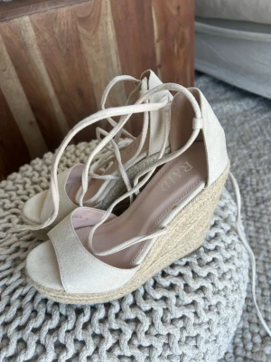 Beiga kilklackade sandaler från RAID - Säljer ett par snygga beiga sandaler från RAID med hög kilklack i flätad jute och öppen tå. Skorna har långa snören att knyta runt ankeln och ovandel i mockaimitation. Perfekta för sommarens alla outfits!