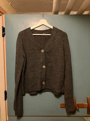 Mörkgrå kofta  - Mysig mörkgrå kofta från 157 med v-ringning och tre stora knappar framtill. Koftan har lång ärm och är stickad i ett mjukt material, perfekt för lager på lager. Enkel och stilren design som passar till det mesta.